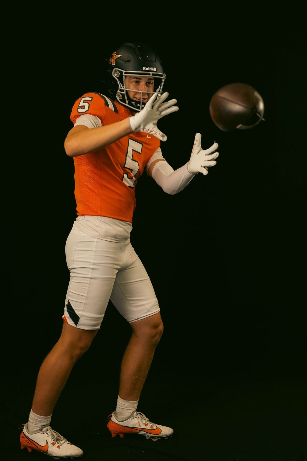 Football player in orange jersey number 5 catches a football against black background.
