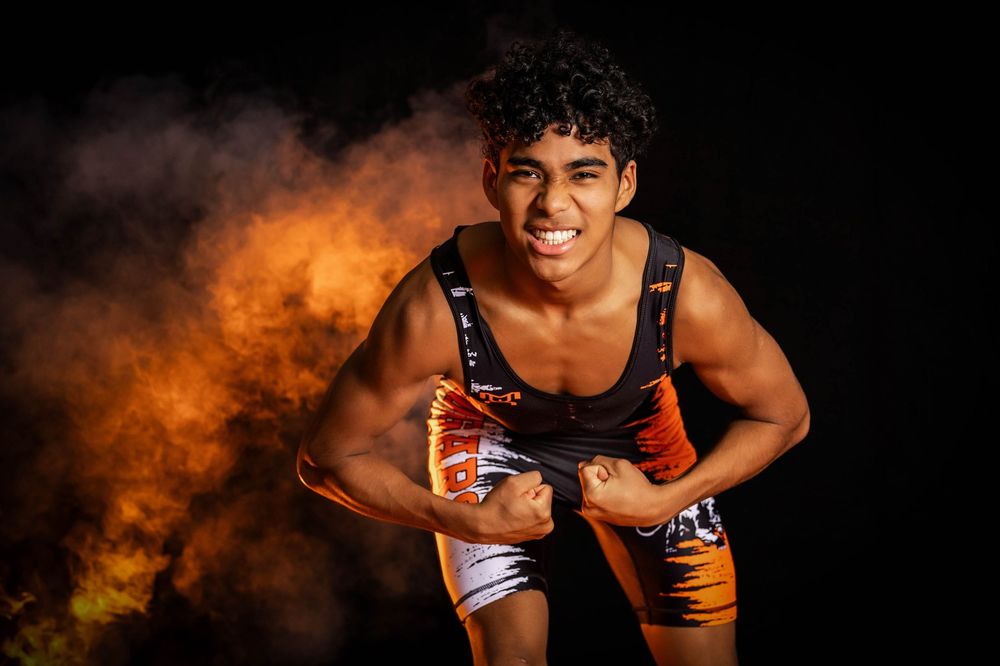 High school wrestler in orange singlet poses in athletic stance with dramatic smoke effect in dark background.