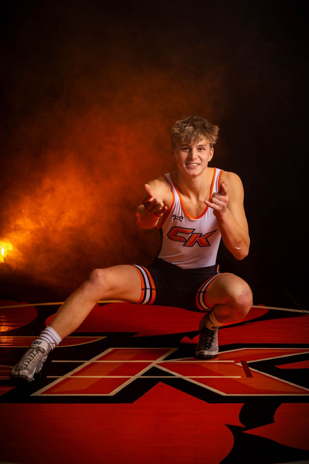 A wrestler in a white and red uniform crouches in a ready stance on a red mat with dramatic orange lighting behind them.