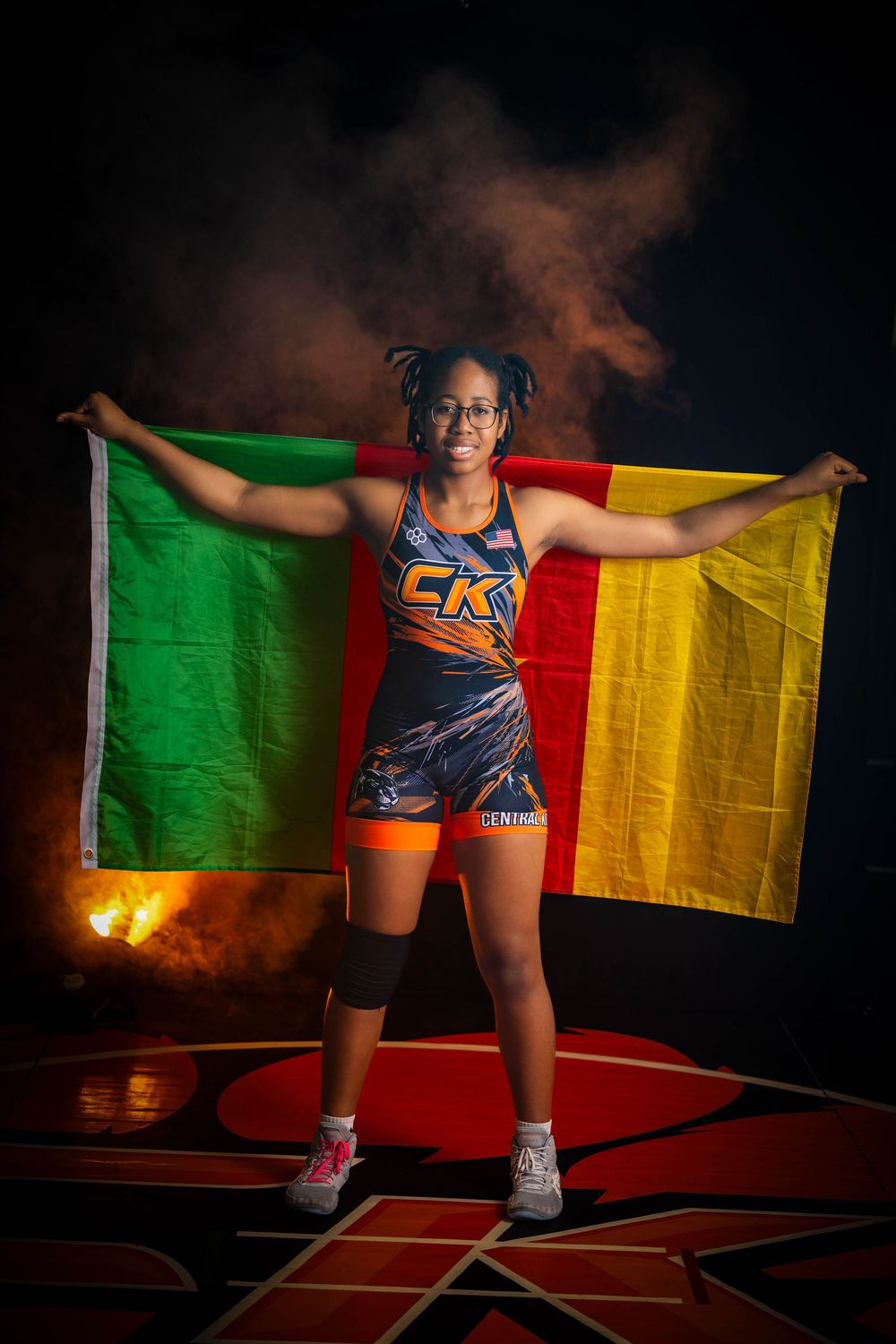 Athlete in blue uniform holds up green, yellow and red striped flag while standing in dramatic lighting and smoke.