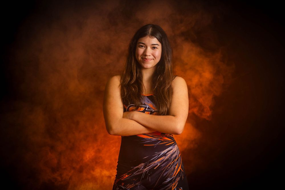 Subject stands confidently with crossed arms against a dramatic dark orange smoky background in a sleeveless dress.
