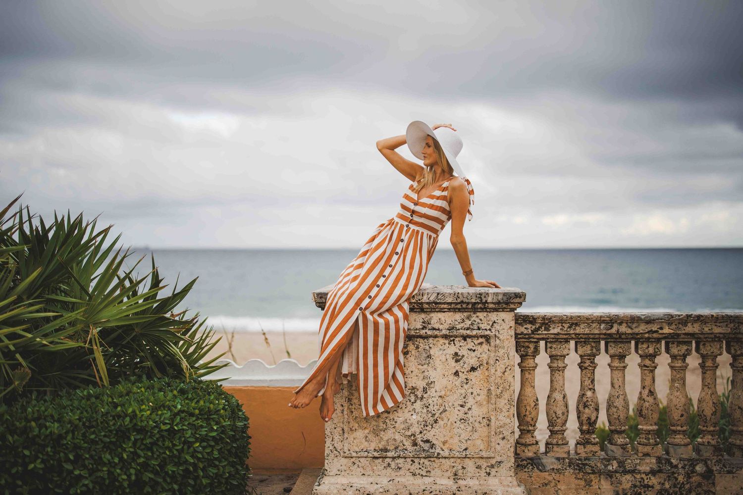 A figure in a striped dress poses dramatically on a stone balustrade overlooking the ocean.