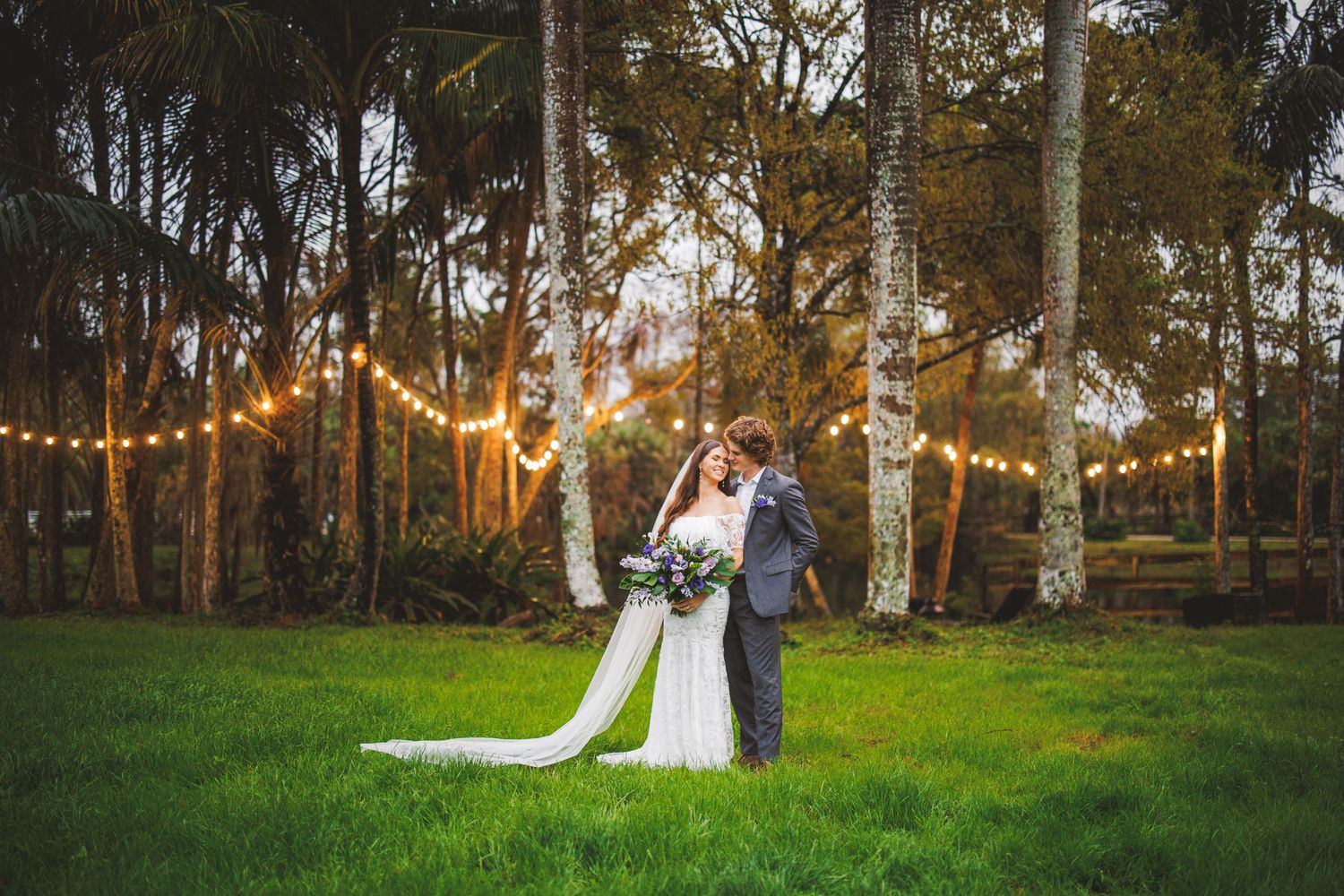 Couple stands beneath illuminated string lights in a woodland setting during sunset wedding ceremony.