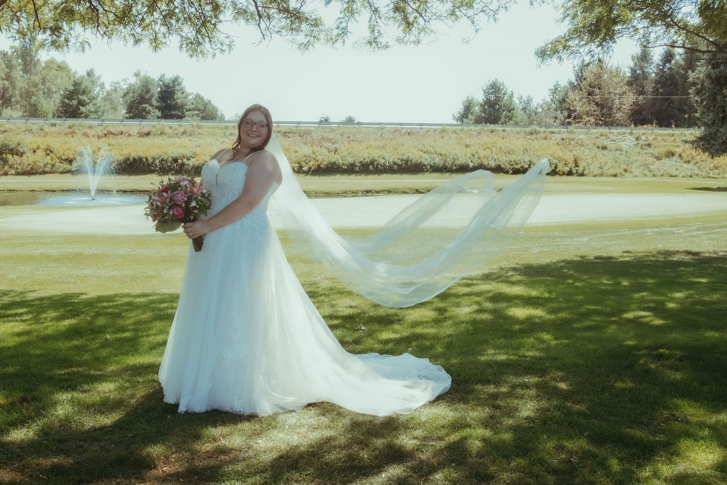A bride in a white gown with flowing veil poses in front of a serene pond on a sunny day.