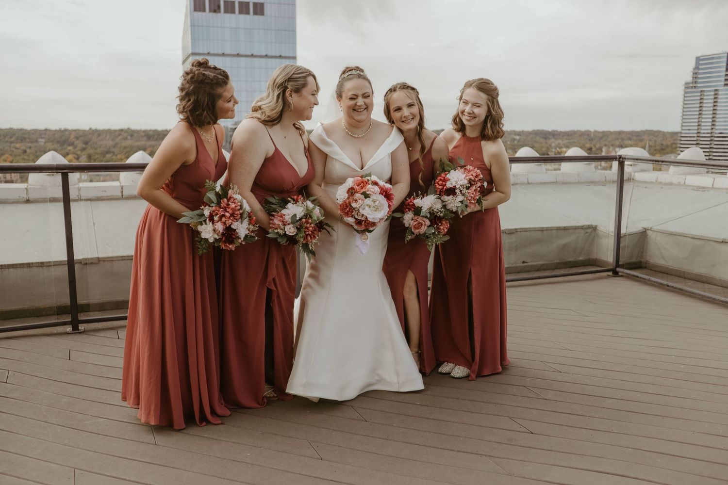A bride in white poses with bridesmaids wearing rust-colored floor-length dresses on a rooftop deck.