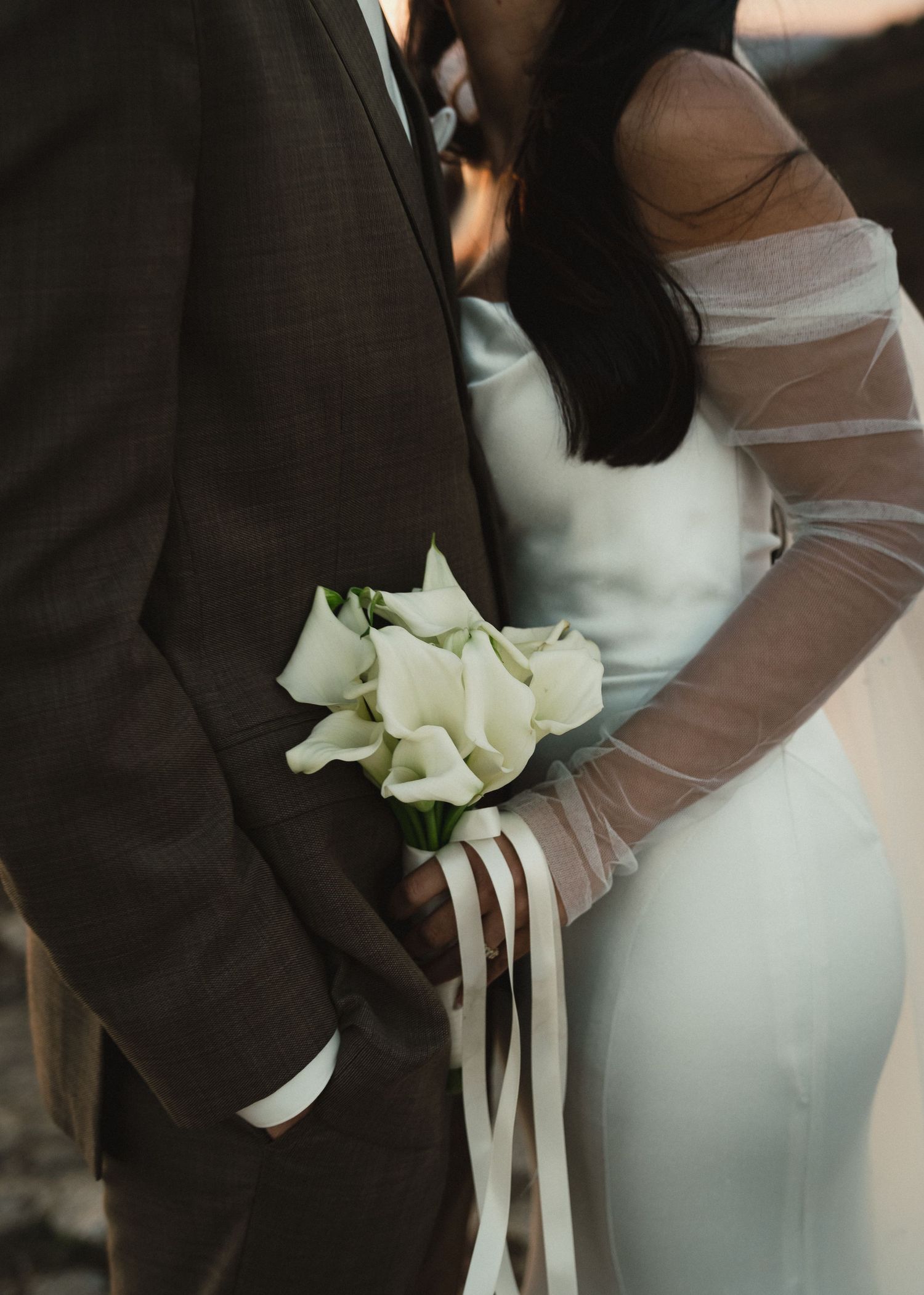 Close-up of a wedding couple with white calla lily boutonniere on brown suit and sheer long sleeve bridal gown.