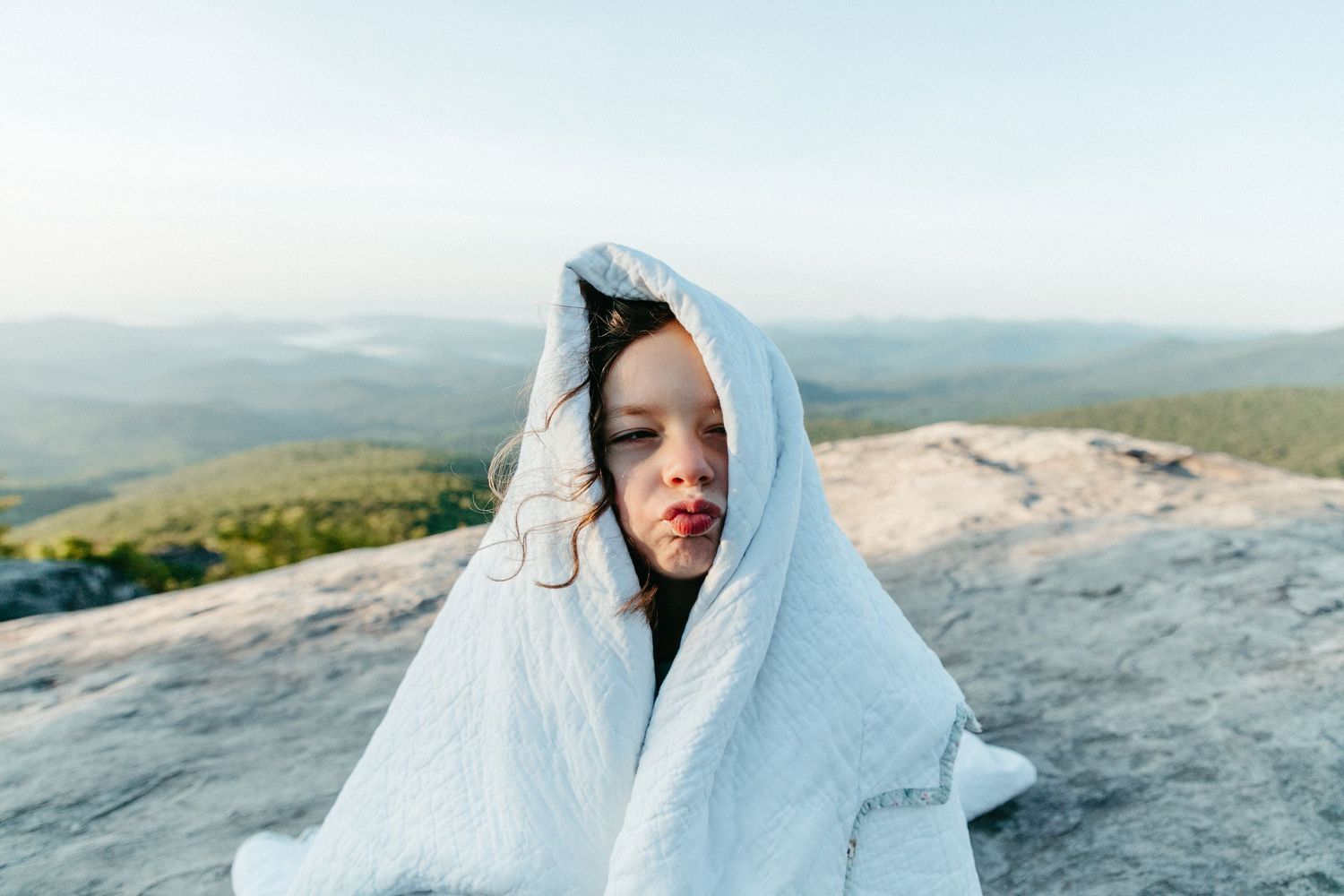 Someone making a silly face wrapped in a light blue blanket stands on a rocky mountain summit overlooking scenic views with windswept hair.