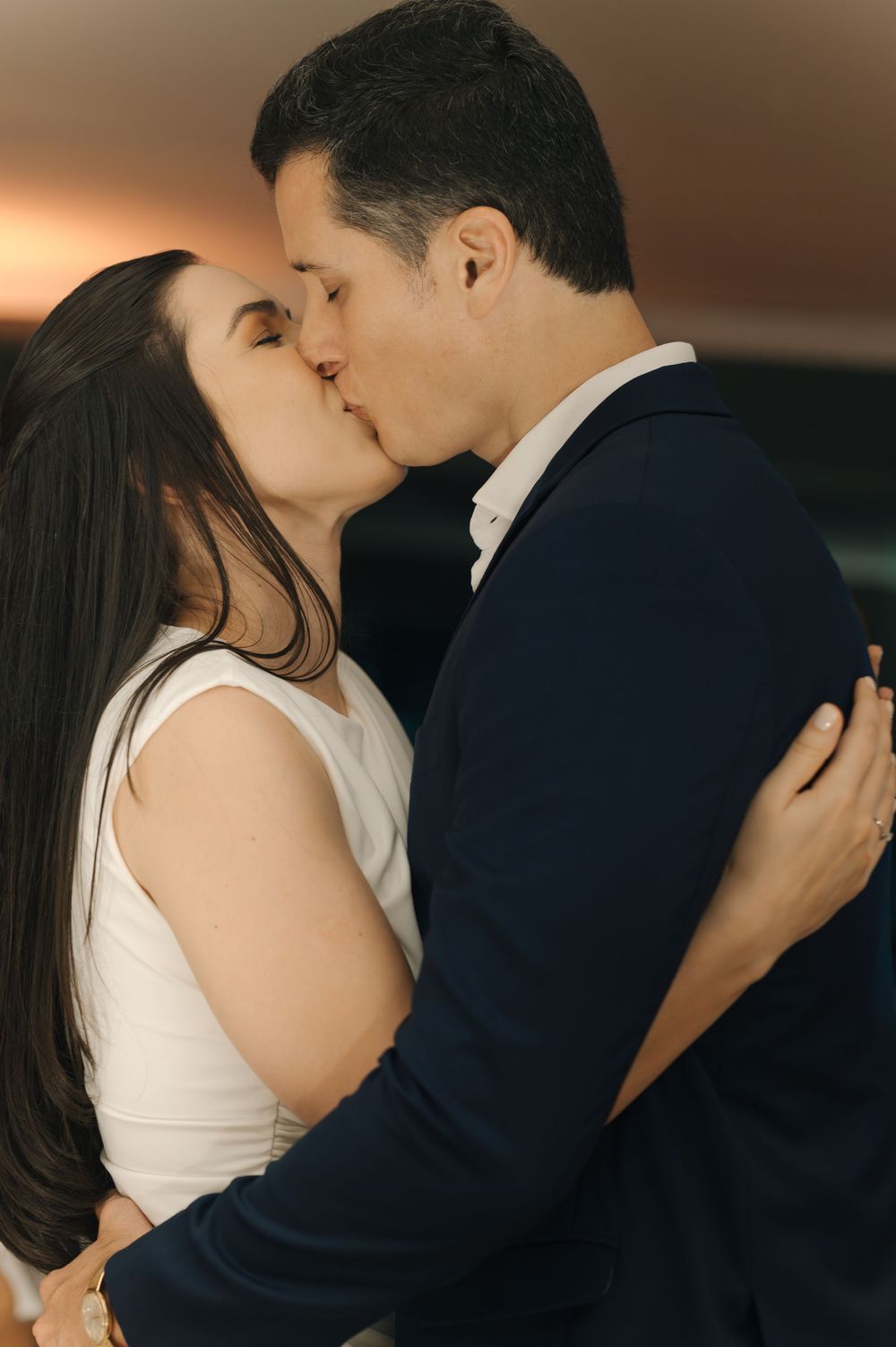 A romantic couple shares an intimate kiss while embracing, with the person in navy suit and white dress clothing.