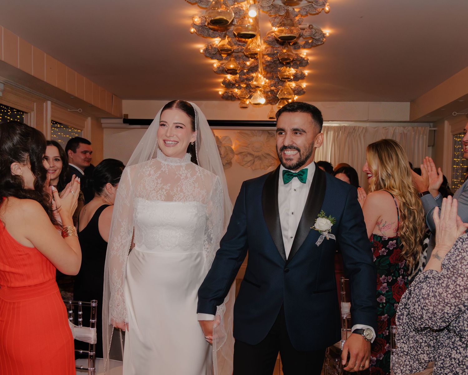 A joyful wedding celebration with newlyweds walking under warm chandelier lighting.