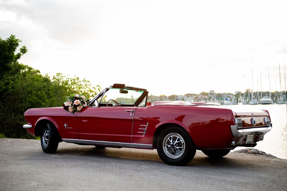 A classic red 1960s Ford Mustang convertible parked near a marina with boats visible in the background.