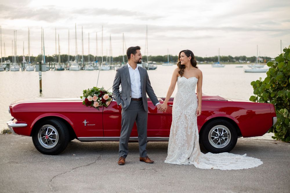 A couple poses next to a classic red Mustang convertible parked at a marina with boats in the background.