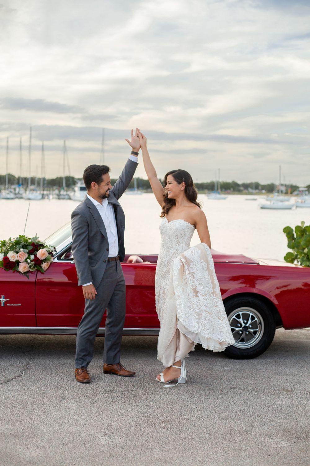 A couple celebrates by a red vintage convertible car at a marina with boats in the background on a cloudy day.