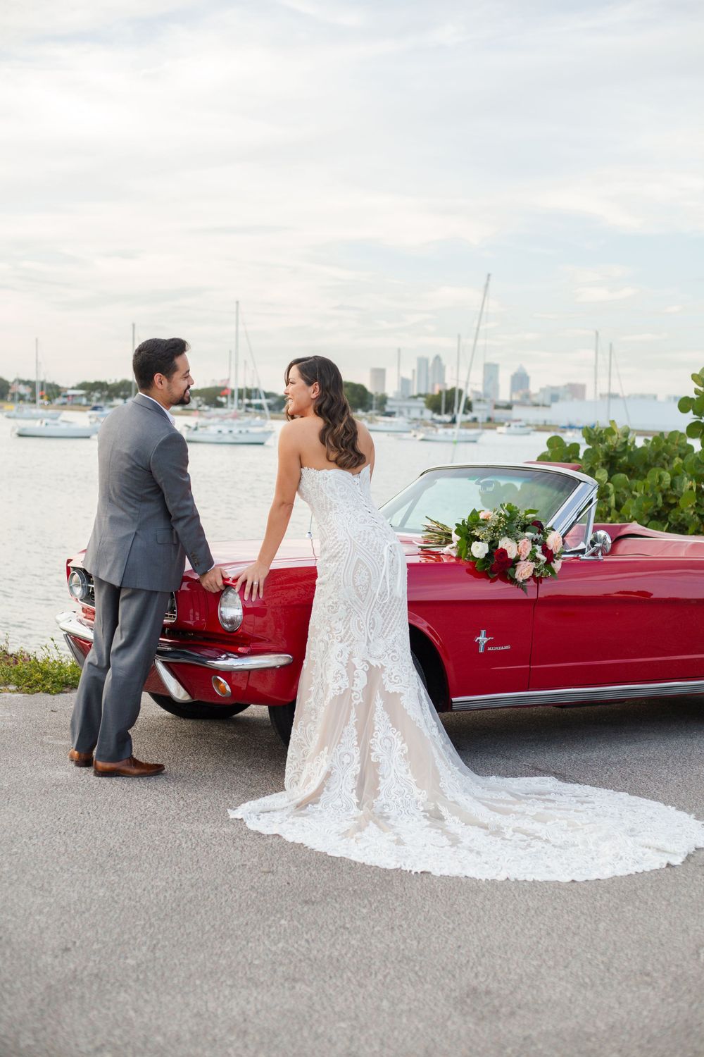 Couple poses with vintage red convertible Mustang at waterfront wedding with city skyline in background.