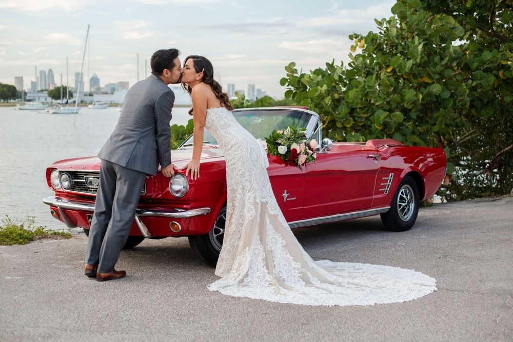 A couple shares a romantic kiss next to a vintage red convertible Mustang with a city skyline visible in the background.