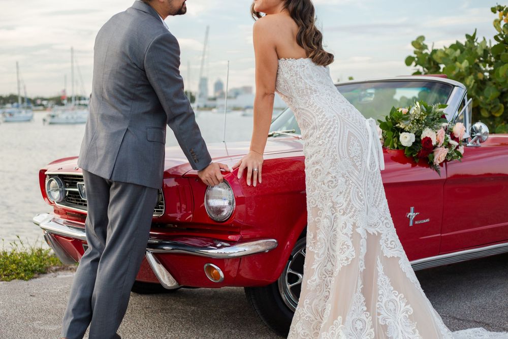 A classic red Ford Mustang convertible serves as backdrop for an elegant wedding photo on a sunny waterfront day.