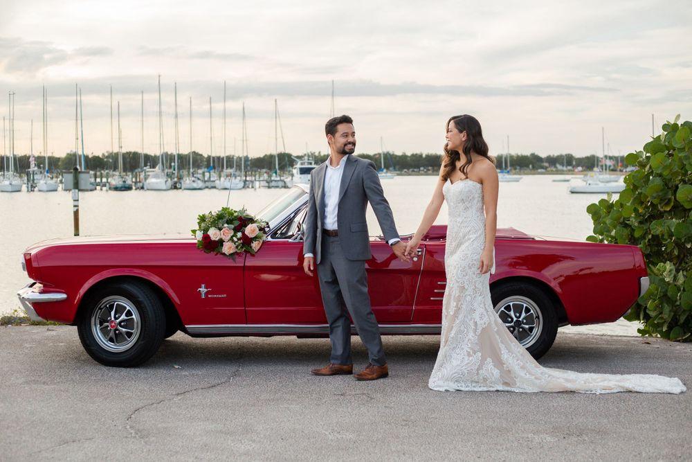 A vintage red Ford Mustang convertible parked at a marina with a couple in wedding attire standing beside it.