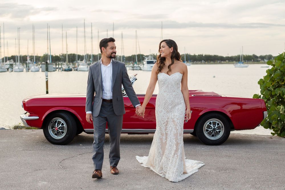 Elegant couple stands next to a classic red vintage car at a marina during sunset, with sailboats in background.
