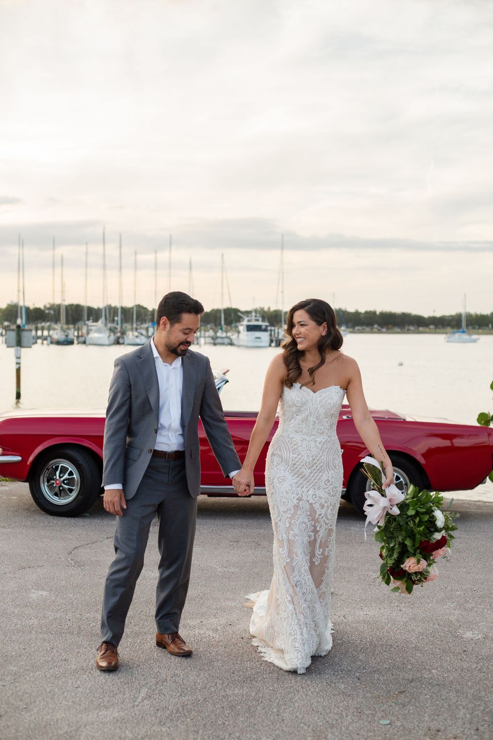 A couple on their wedding day walking along a marina dock with a classic red convertible car in the background.
