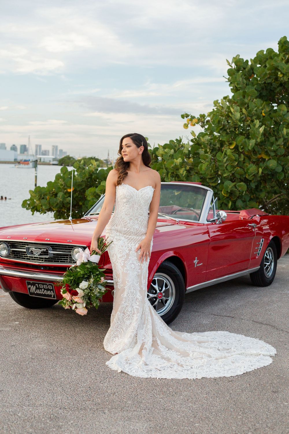 Classic red Ford Mustang convertible with bridal bouquet and elegant wedding gown against city skyline backdrop.