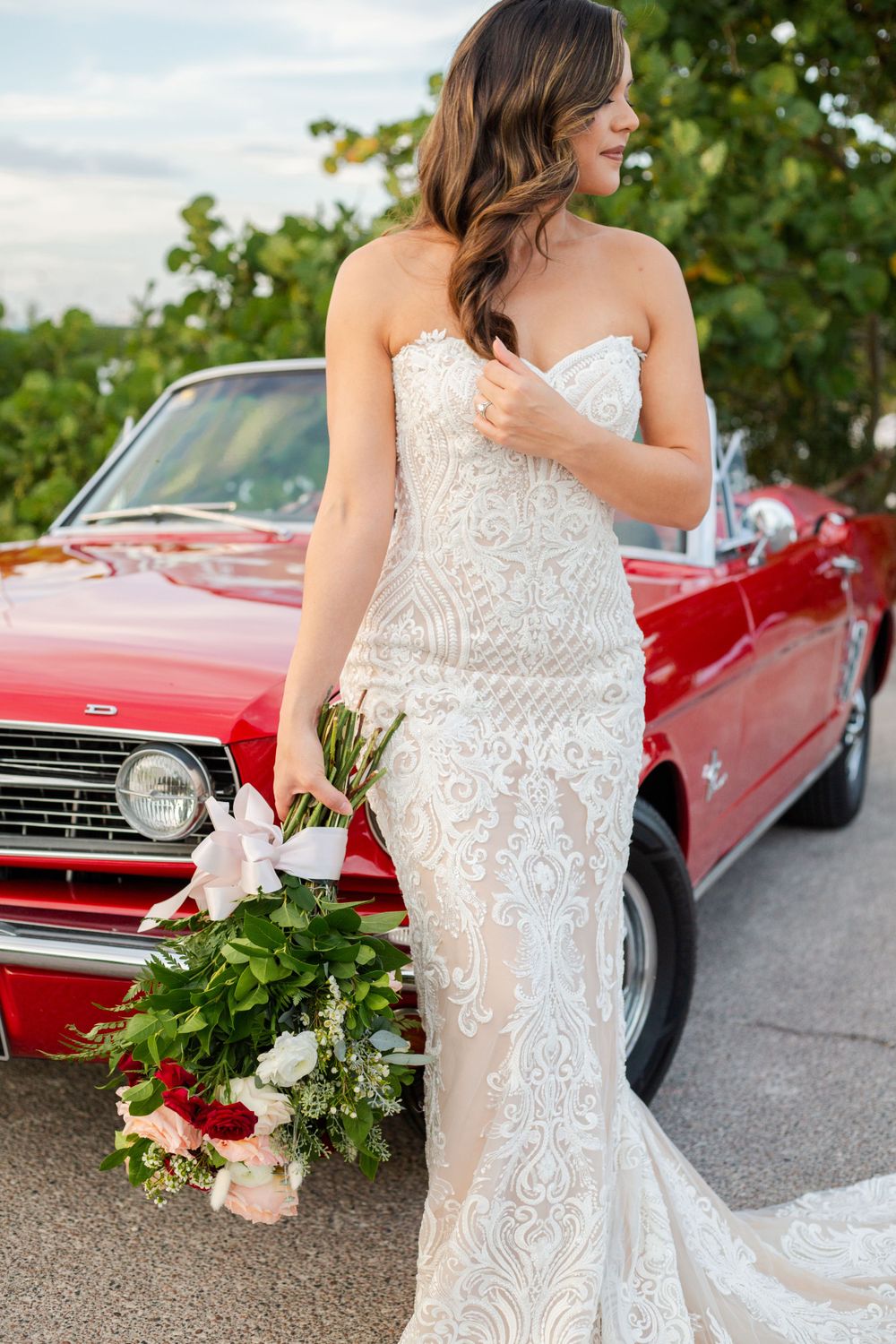 Bride in intricate lace wedding gown holds bouquet while standing next to vintage red convertible car.