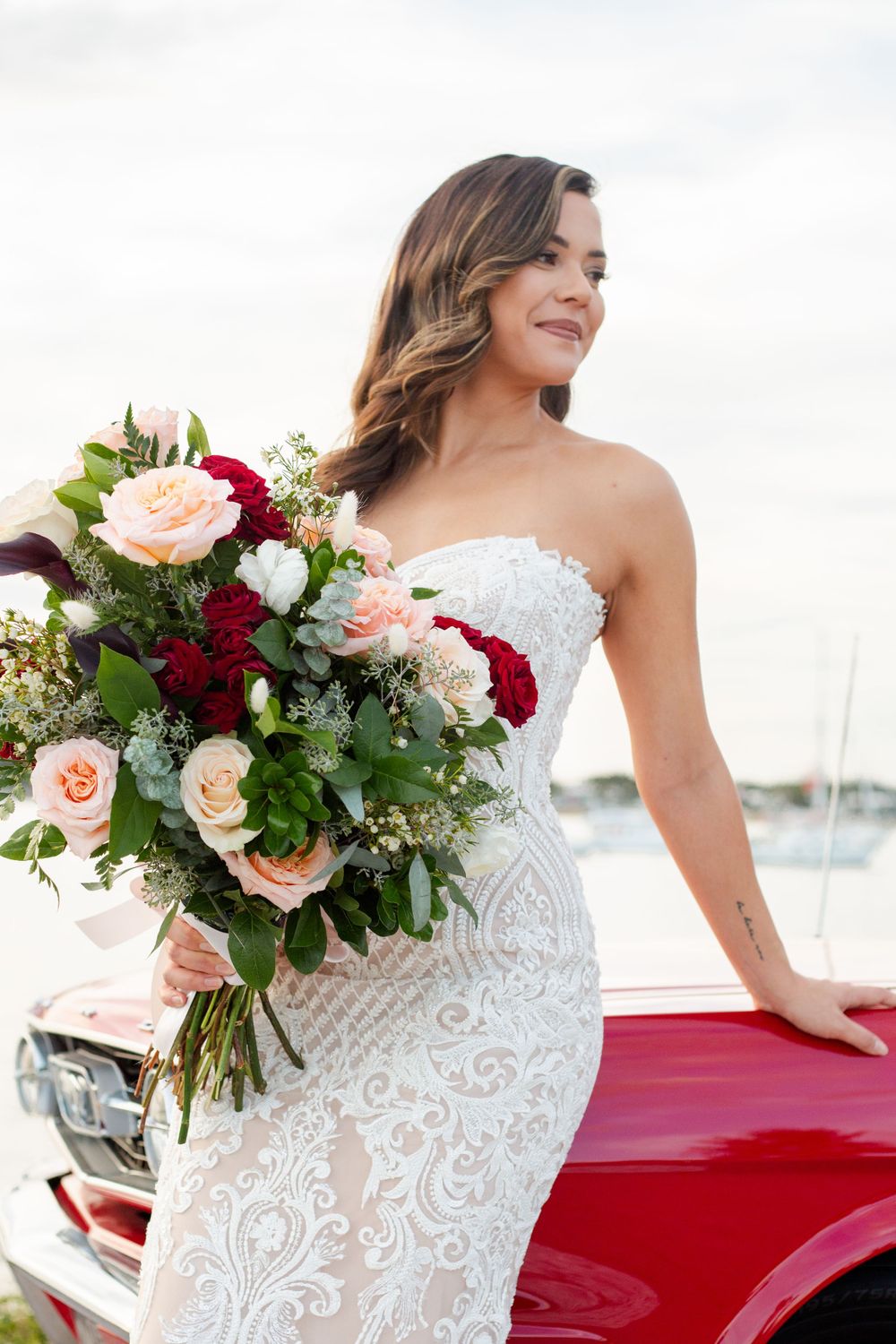 A bride in a lace wedding gown holds a peach and burgundy floral bouquet while posing against a red vintage car near a marina.