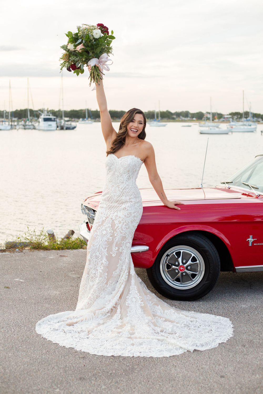 A bride in a lace wedding gown poses with a bouquet next to a classic red Mustang car at a marina.