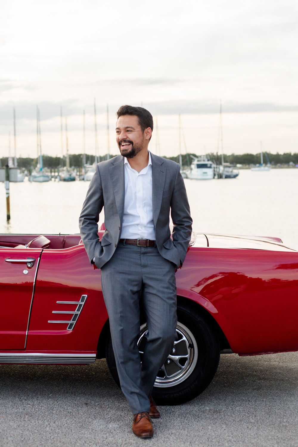 A person in a gray suit leans against a classic red convertible car parked at a marina with boats in the background.