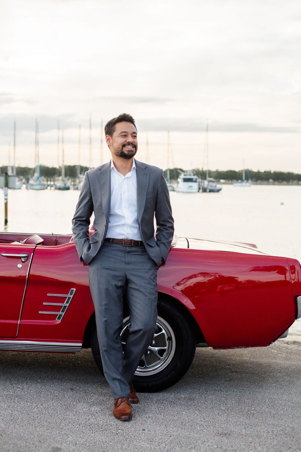 Well-dressed person in gray suit leaning against a classic red convertible car near a marina with boats in background.