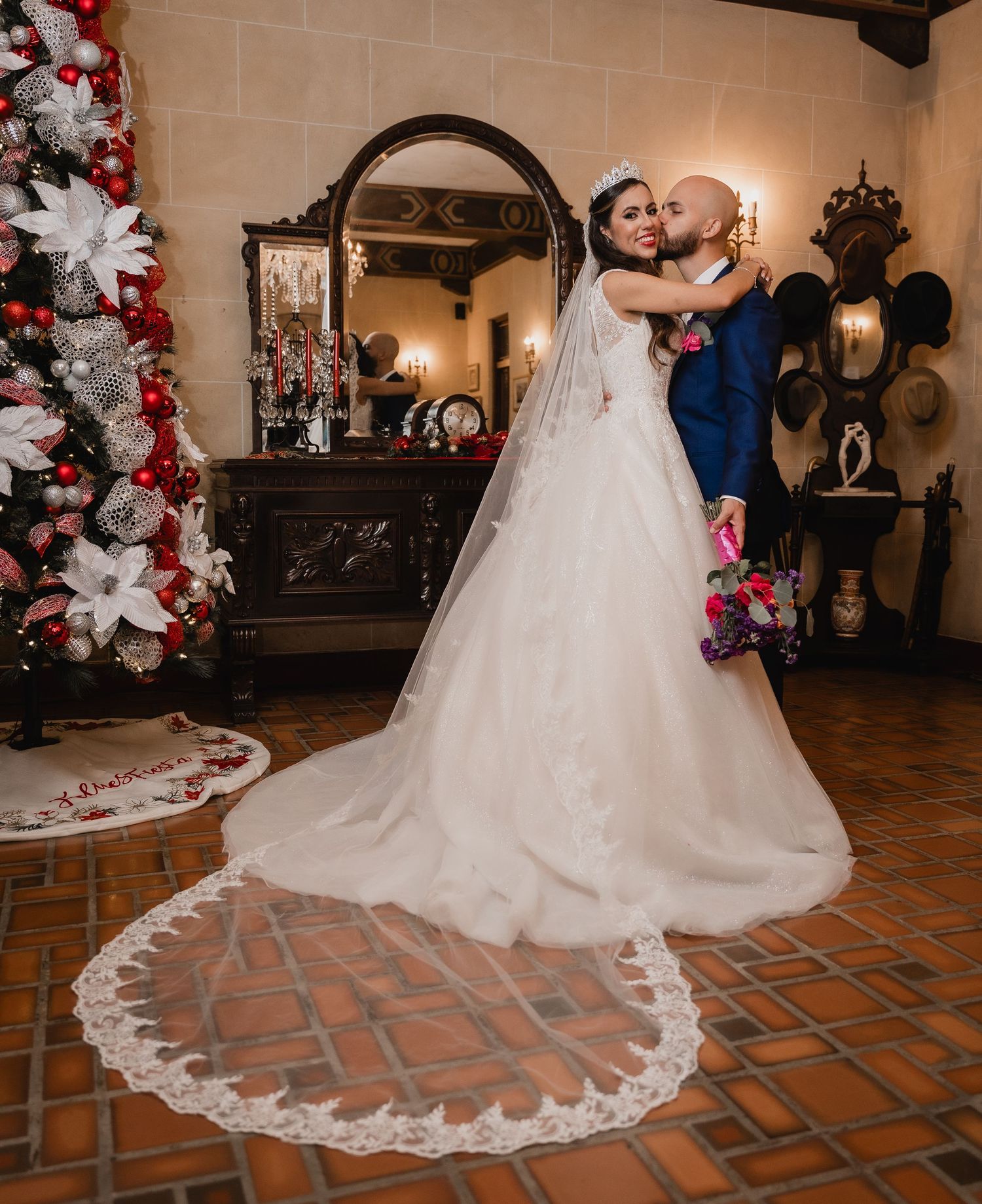 A couple shares a romantic moment beneath an ornate mirror, with a white floral Christmas tree and brick floor in view.