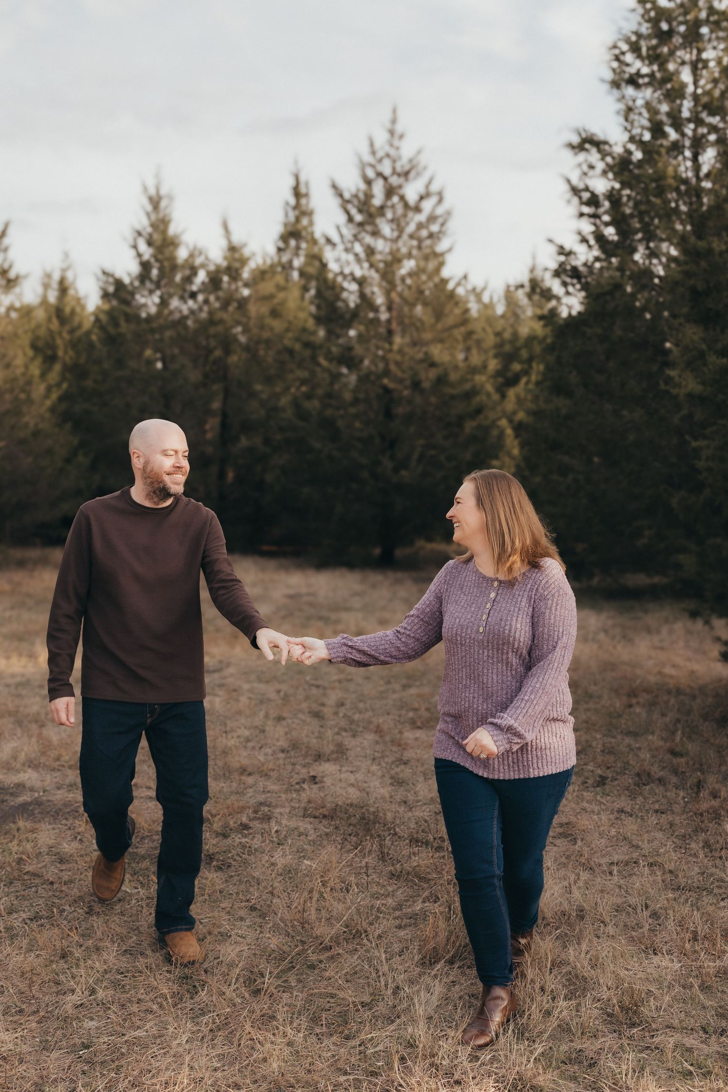 Couple in cozy sweaters holding hands while walking through a field with pine trees.
