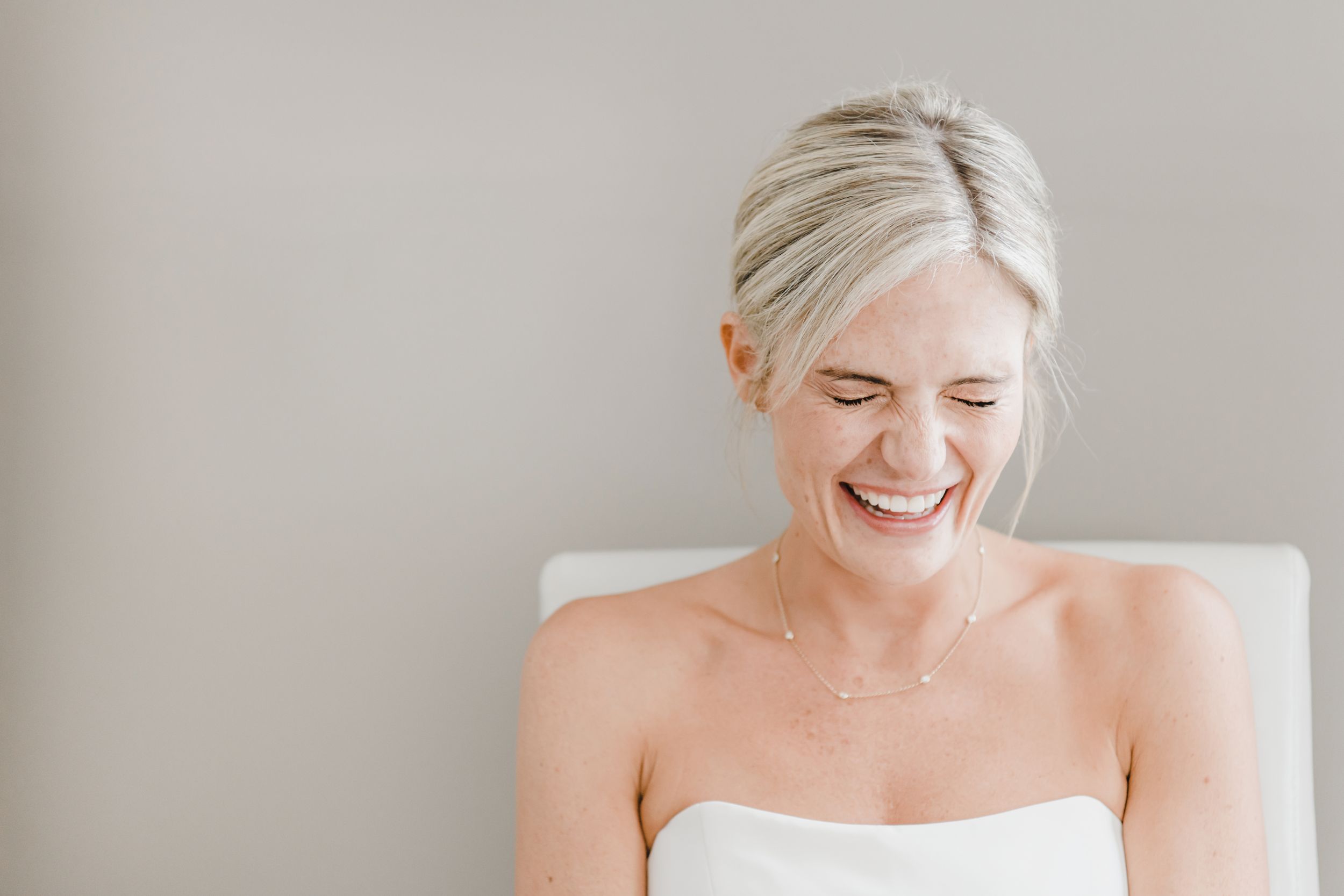 A glowing bride wearing a white strapless wedding dress laughs joyfully with natural lighting in the background.
