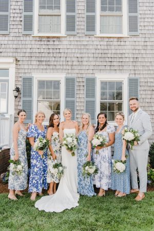 Wedding party in coordinating blue and white floral dresses poses together on grass in front of shingled house.