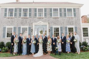 Wedding party in blue floral dresses and dark suits pose together outside a classic white New England style house.
