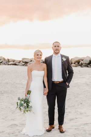 Wedding couple stands hand in hand on a sandy beach at sunset with rocks and ocean in the background.