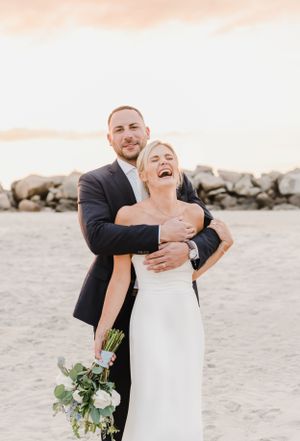 Joyful wedding couple shares a candid moment laughing together on a beach with rocks and sunset in background.