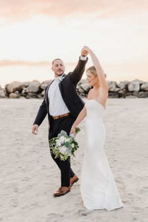 A couple dancing joyfully on a beach at sunset with rocky breakwater in background and white wedding bouquet.