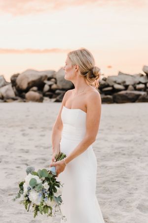 A bride in a sleek strapless white gown holds a lush white and green bouquet while standing on a sandy beach at sunset.