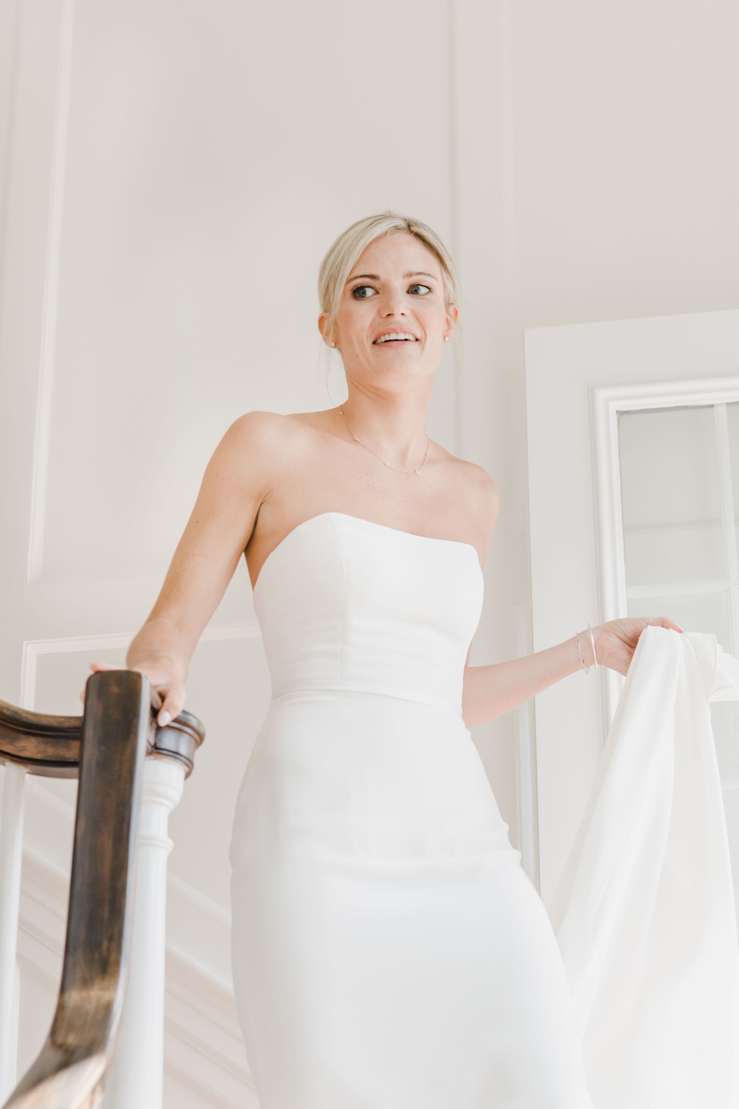 A person in a strapless white wedding dress stands on a staircase while smiling and holding fabric.
