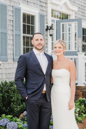 A couple stands together on a white porch with light blue shutters and hydrangeas in the garden.