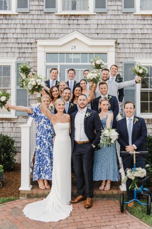A wedding party gathered on the front steps of a shingled beach house decorated with white floral arrangements.