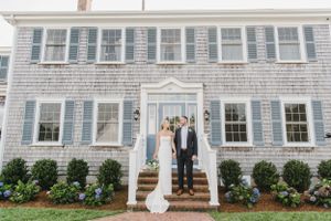 A couple stands under a white archway entrance of a classic Cape Cod style shingled house with blue shutters and hydrangeas.