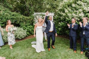 Wedding party celebrates outdoors in a garden setting with lush greenery and floral arch backdrop.
