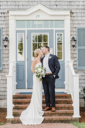 Elegant couple in wedding attire share a romantic moment on brick steps in front of a classic white door with blue accents.