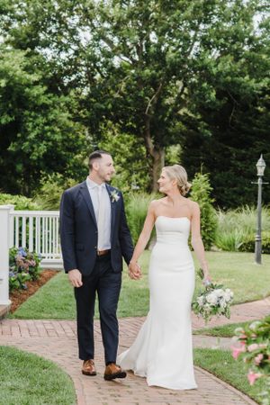 A couple in wedding attire walks hand in hand along a brick path bordered by gardens and white railing on a summer day.