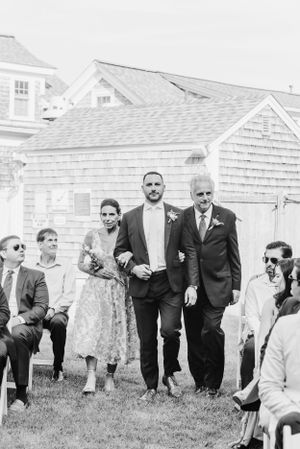 A wedding party in formal attire walks across a grassy lawn with white shingled buildings in the background.