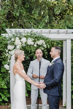A wedding ceremony takes place under a white pergola decorated with lush greenery and white floral arrangements.