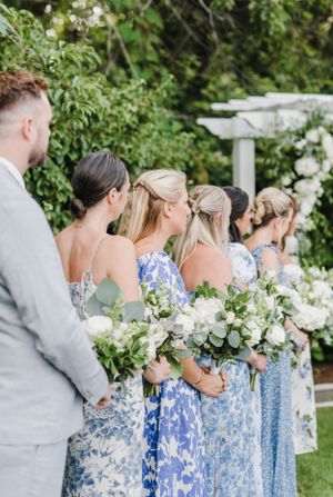 Wedding party wears coordinated blue floral dresses while holding white bouquets in an outdoor garden setting.