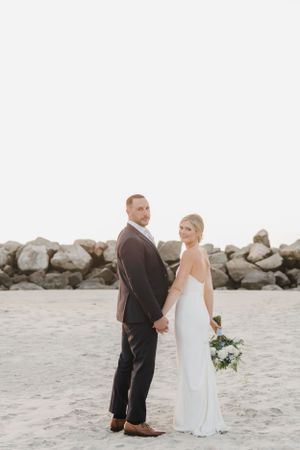 A wedding couple stands holding hands on a sandy beach with a rocky breakwall in the background during sunset.