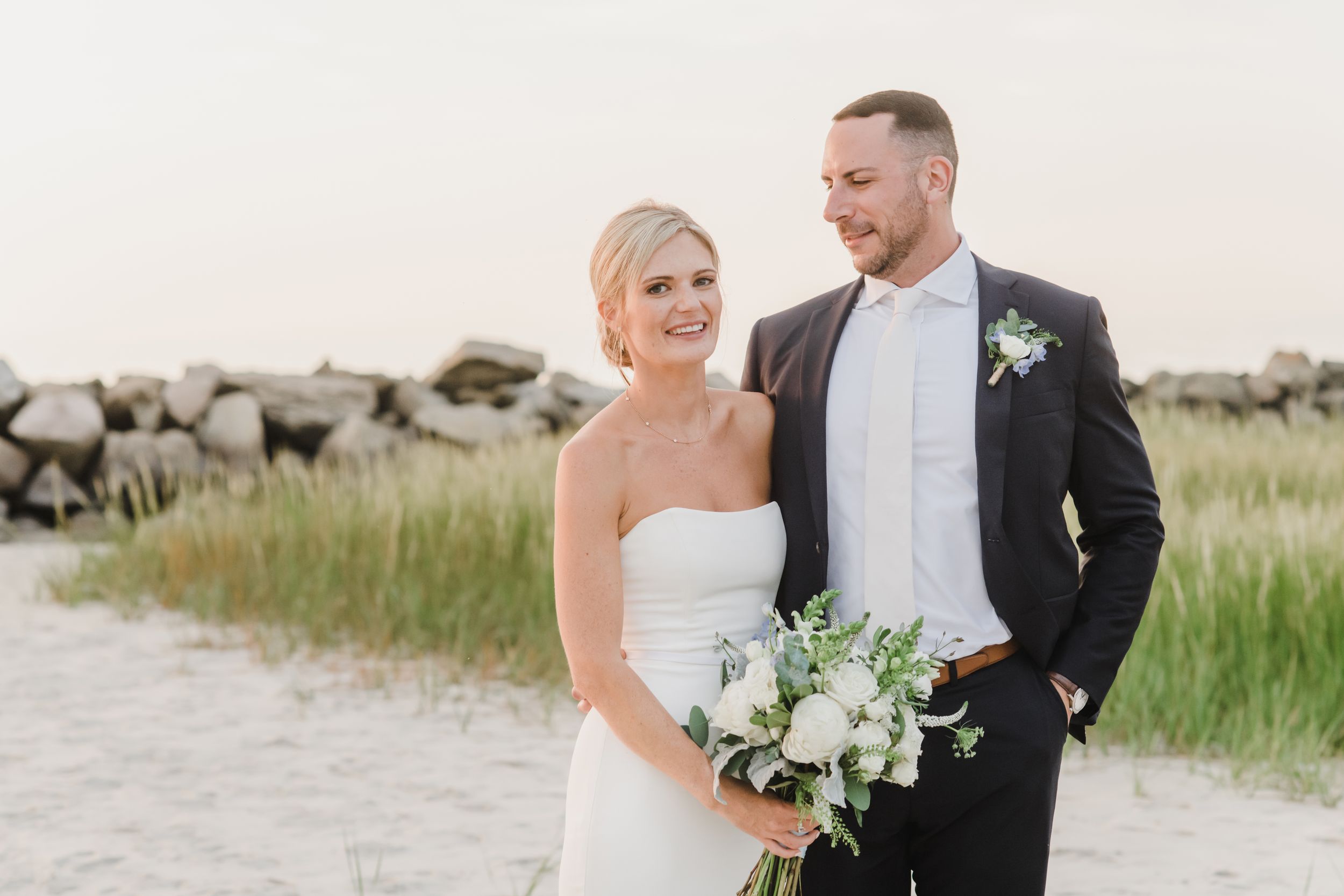 A couple stands together on a beach path with rock formations and grass in the background.