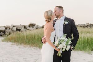 A couple in wedding attire embraces on a beach path with a bouquet of white flowers against a coastal landscape.